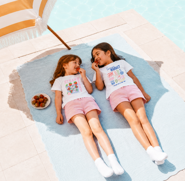 Two children lying on a towel poolside under a striped beach umbrella eating strawberries